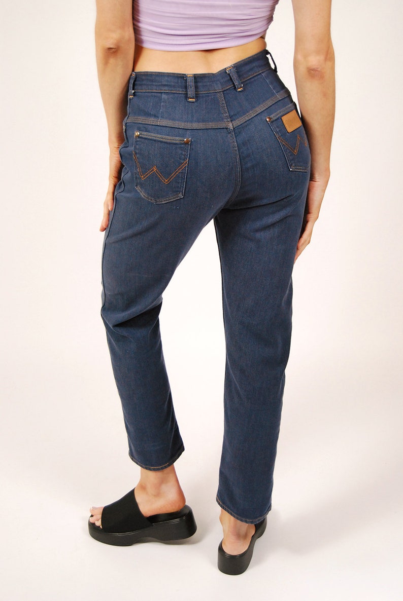 May include: Dark blue denim jeans with a high waist and straight leg. Features include orange stitching, a brown leather patch, and the Wrangler logo on the back pocket. The jeans are worn with black platform sandals.