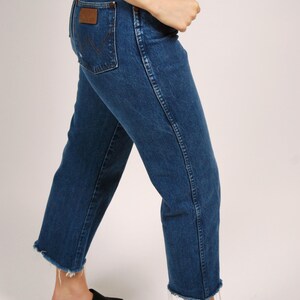 Op de afbeelding: Blauwe denim jeans met een klassieke pasvorm en een rauwe zoom. De jeans hebben een bruine leren patch op de achterzak en een rood logo. De persoon draagt ​​zwarte plateausandalen. Een casual, alledaagse stijl.