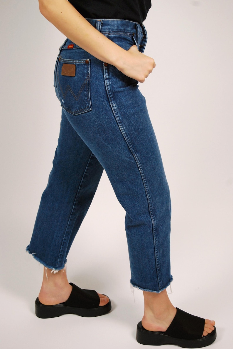 Op de afbeelding: Blauwe denim jeans met een klassieke pasvorm en een rauwe zoom. De jeans hebben een bruine leren patch op de achterzak en een rood logo. De persoon draagt ​​zwarte plateausandalen. Een casual, alledaagse stijl.