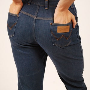 May include: Dark blue denim jeans with orange stitching details. Features include back pockets with a signature "W" stitch and a brown leather patch with the "Wrangler" logo. Classic five-pocket design.