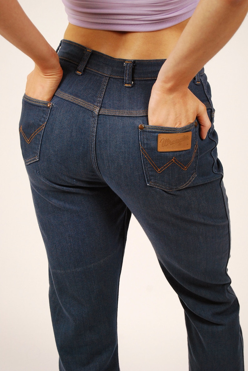 May include: Dark blue denim jeans with orange stitching details. Features include back pockets with a signature "W" stitch and a brown leather patch with the "Wrangler" logo. Classic five-pocket design.