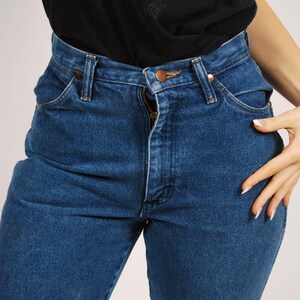 Op de afbeelding: Close-up van een blauwe spijkerbroek met een rits en een koperkleurige knoop. De jeans wordt gedragen met een zwart t-shirt met de woorden "THE LION KING" en "THE BROADWAY MUSICAL" in het geel.