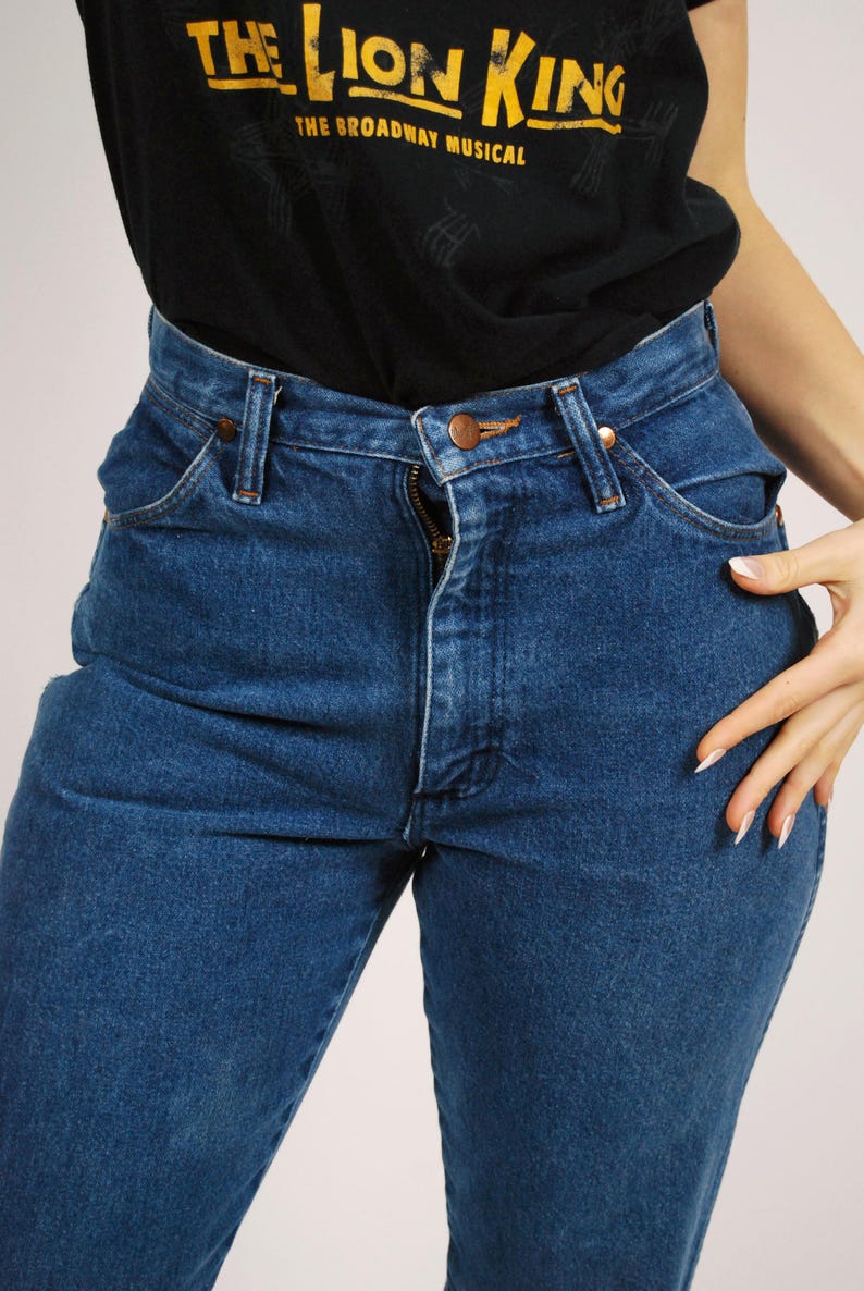 Op de afbeelding: Close-up van een blauwe spijkerbroek met een rits en een koperkleurige knoop. De jeans wordt gedragen met een zwart t-shirt met de woorden "THE LION KING" en "THE BROADWAY MUSICAL" in het geel.