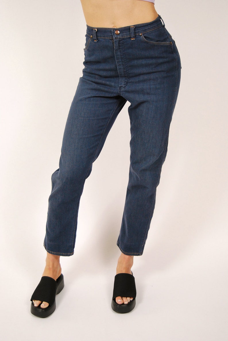 May include: Dark wash denim jeans with a high waist and straight leg. The jeans have a classic five-pocket design and a button and zipper closure. The model is wearing black platform sandals.