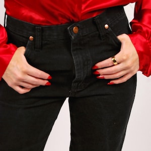 May include: Black denim jeans with copper-colored rivets and a button, paired with a bright red satin blouse. The blouse has long sleeves. The person is wearing a gold ring and has red nail polish.