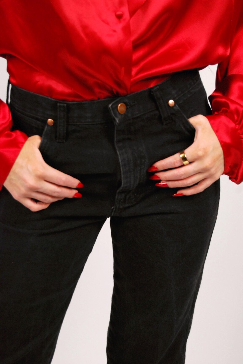 May include: Black denim jeans with copper-colored rivets and a button, paired with a bright red satin blouse. The blouse has long sleeves. The person is wearing a gold ring and has red nail polish.