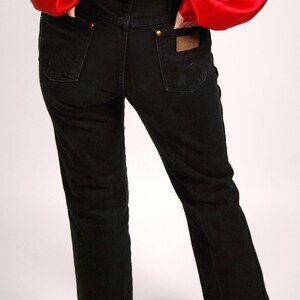 May include: Black denim jeans with a classic straight leg fit. The jeans feature a brown leather patch on the back pocket and gold-colored rivets. The person is wearing a red satin top.