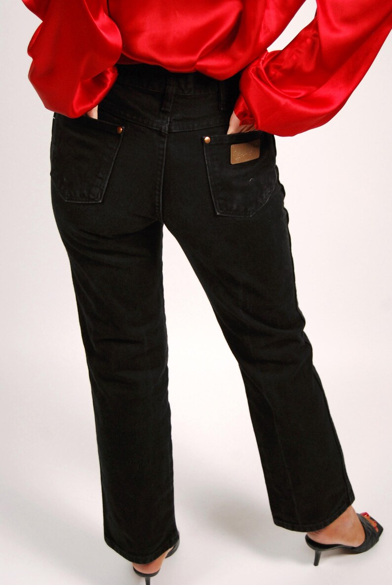 May include: Black denim jeans with a classic straight leg fit. The jeans feature a brown leather patch on the back pocket and gold-colored rivets. The person is wearing a red satin top.