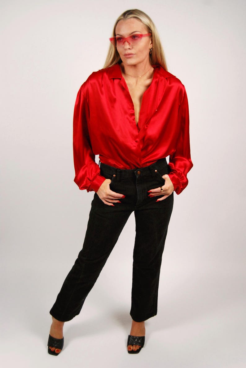 May include: A red satin blouse with long sleeves and a collared neckline paired with black denim jeans. The outfit is completed with black open-toe heels. The model is wearing pink-tinted sunglasses.