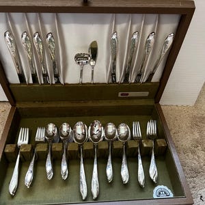 May include: A silver-toned flatware set arranged in a dark brown wooden storage chest. The set includes various forks, spoons, and knives with ornate handles, displayed against a white satin-lined lid and a dark green felt-lined base.