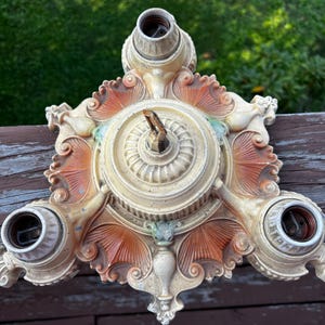 May include: A three-light ceiling fixture base featuring a cream-colored finish with orange-toned accents. The metal structure displays intricate molded details, including fan-shaped motifs and scrollwork, with three circular light sockets.