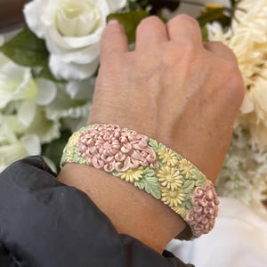 May include: A floral bracelet featuring a repeating pattern of pink, yellow, and green flowers. The bracelet is worn on a wrist and appears to be made of a hard material. The flowers are intricately detailed.