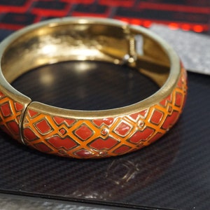 May include: A gold bangle bracelet with a red and orange geometric pattern.
