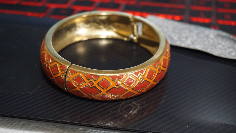 May include: A gold bangle bracelet with a red and orange geometric pattern.