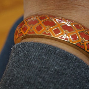 May include: A gold bangle bracelet with a red and orange geometric pattern. The bracelet is on a person's wrist.