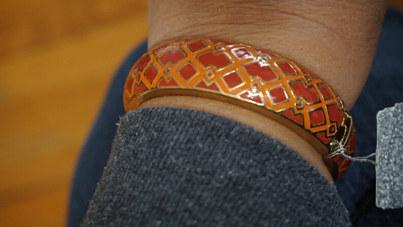 May include: A gold bangle bracelet with a red and orange geometric pattern. The bracelet is on a person's wrist.