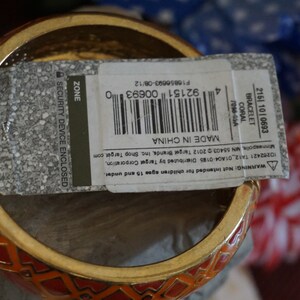 May include: A red and gold bangle bracelet with a geometric pattern. The bracelet has a tag attached that reads "Made in China" and includes a barcode. The tag also says "Security Device Enclosed".