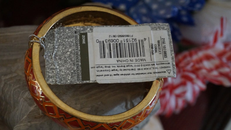 May include: A red and gold bangle bracelet with a geometric pattern. The bracelet has a tag attached that reads "Made in China" and includes a barcode. The tag also says "Security Device Enclosed".