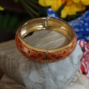 May include: A gold bangle bracelet with a red and orange geometric pattern. The bracelet is open and has a hinged clasp.