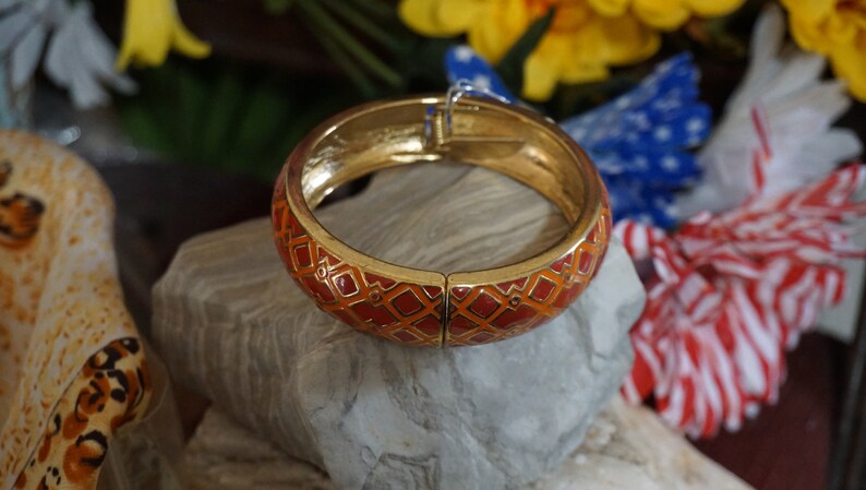 May include: A gold bangle bracelet with a red and orange geometric pattern. The bracelet is open and has a hinged clasp.