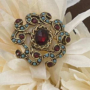 May include: A circular gold-tone brooch featuring a central oval deep red gemstone surrounded by a decorative frame of swirling metalwork. The design is accented with small, round, light blue beads and additional small, round, deep red gemstones.