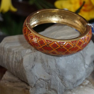 May include: A gold bangle bracelet with a red and orange geometric pattern. The bracelet is sitting on a gray rock.