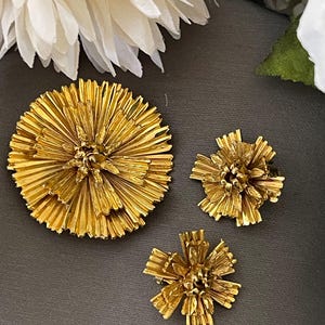 K&ouml;nnte beinhalten: Drei goldfarbene Broschen in Form von Blumen. Die gr&ouml;&szlig;te Brosche ist eine runde Blume mit mehreren Bl&uuml;tenblattschichten. Die beiden kleineren Broschen sind kleinere, kompaktere Blumen mit einem &auml;hnlichen Design.