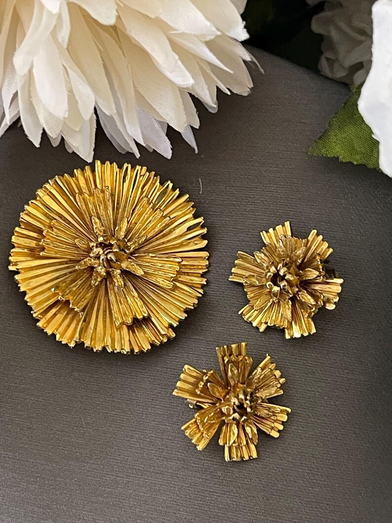 K&ouml;nnte beinhalten: Drei goldfarbene Broschen in Form von Blumen. Die gr&ouml;&szlig;te Brosche ist eine runde Blume mit mehreren Bl&uuml;tenblattschichten. Die beiden kleineren Broschen sind kleinere, kompaktere Blumen mit einem &auml;hnlichen Design.