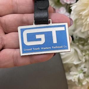 May include: A vintage GT keychain with a blue background and white lettering. The keychain has a black leather strap and the text "Grand Trunk Western Railroad Co". The metal frame has a silver finish.