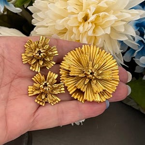K&ouml;nnte beinhalten: Ein Set aus drei goldfarbenen Broschen in Blumenform. Die gr&ouml;&szlig;te Brosche befindet sich in der Mitte, die beiden kleineren Broschen befinden sich auf beiden Seiten. Die Broschen bestehen aus d&uuml;nnem, &uuml;bereinanderliegendem Metall, das einen strukturierten, dreidimensionalen Look erzeugt.