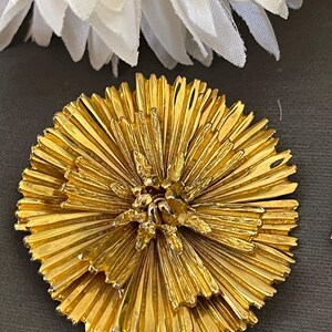 K&ouml;nnte beinhalten: Eine goldfarbene Brosche in Form einer Blume mit einem mehrschichtigen, plissierten Design. Die Blume hat eine Mitte mit einem kleineren, detaillierteren Blumendesign.