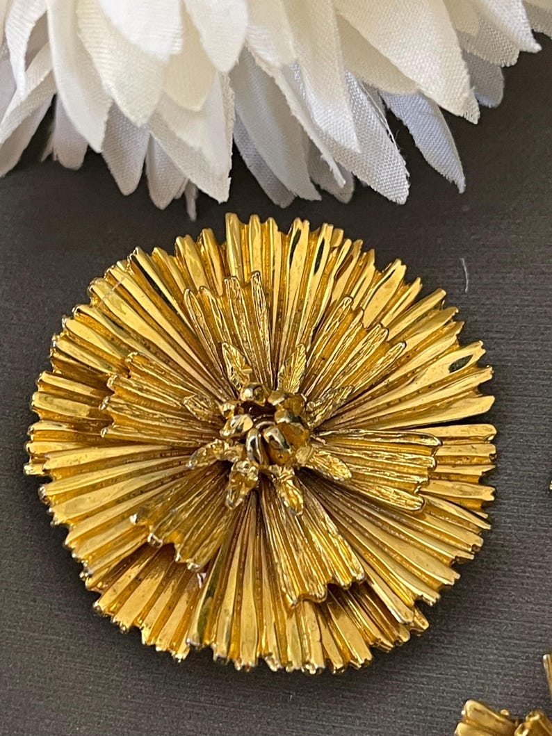 K&ouml;nnte beinhalten: Eine goldfarbene Brosche in Form einer Blume mit einem mehrschichtigen, plissierten Design. Die Blume hat eine Mitte mit einem kleineren, detaillierteren Blumendesign.