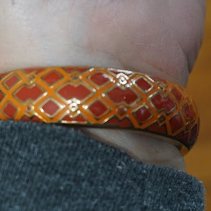 May include: A gold bangle bracelet with a red and orange geometric pattern.