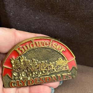 May include: A gold-colored belt buckle with a red and white Budweiser Clydesdales logo. The buckle is shaped like a half-circle and features a raised image of the Clydesdales pulling a wagon.