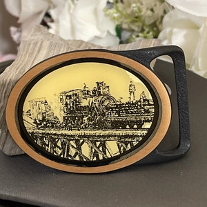 May include: A gold-toned belt buckle with a black and white image of a train engine and a bridge. The buckle is attached to a black leather belt.