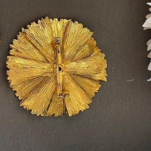 K&ouml;nnte beinhalten: Eine goldfarbene Brosche mit einem runden Design, das ein strahlendes Muster aus d&uuml;nnen, strukturierten Metallstreifen aufweist. Die Brosche hat eine Nadel und einen Verschluss auf der R&uuml;ckseite.