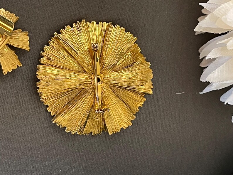 K&ouml;nnte beinhalten: Eine goldfarbene Brosche mit einem runden Design, das ein strahlendes Muster aus d&uuml;nnen, strukturierten Metallstreifen aufweist. Die Brosche hat eine Nadel und einen Verschluss auf der R&uuml;ckseite.