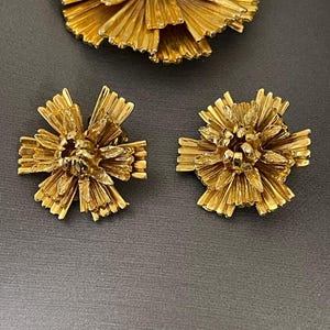 K&ouml;nnte beinhalten: Ein Set aus goldfarbenem Schmuck mit einer gro&szlig;en Blumenbrosche und zwei passenden Ohrringen. Das Blumendesign besteht aus d&uuml;nnen, filigranen Metallteilen, die einen strukturierten, dreidimensionalen Look erzeugen.