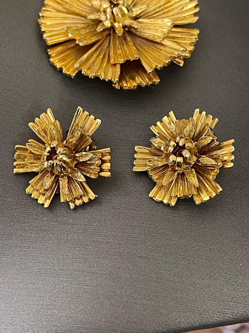 K&ouml;nnte beinhalten: Ein Set aus goldfarbenem Schmuck mit einer gro&szlig;en Blumenbrosche und zwei passenden Ohrringen. Das Blumendesign besteht aus d&uuml;nnen, filigranen Metallteilen, die einen strukturierten, dreidimensionalen Look erzeugen.