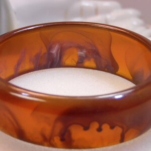 May include: A brown and amber colored plastic bangle bracelet with a swirling pattern. The bracelet is smooth and shiny.