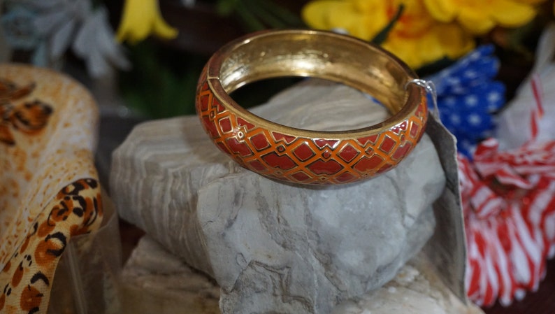 May include: A gold bangle bracelet with a red and orange geometric pattern. The bracelet is sitting on a gray rock.