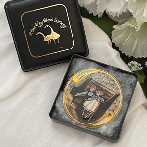 May include: A round brooch with a gold-colored trim, featuring a painted scene of two figures on a bench. The brooch is in a black box with the text "P. Buckley Moss Society" and a gold-colored goose logo.