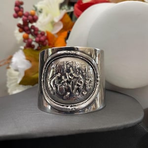 May include: A wide silver cuff bracelet featuring a central circular medallion with a detailed embossed relief scene of figures in a forest setting, framed by a beaded border and a polished, smooth metallic band finish.