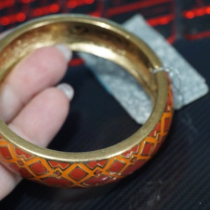 May include: A gold bangle bracelet with an orange and gold geometric pattern. The bracelet is open and has a smooth, polished finish.
