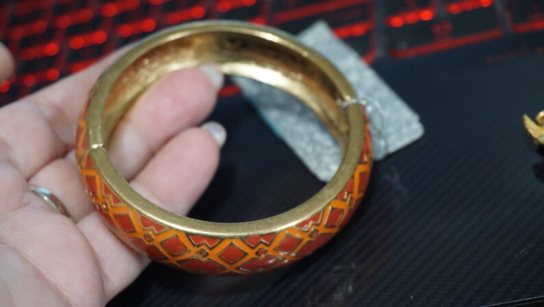 May include: A gold bangle bracelet with an orange and gold geometric pattern. The bracelet is open and has a smooth, polished finish.