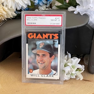May include: A graded baseball card featuring Will Clark, a former first baseman for the San Francisco Giants. The card is from the 1986 Topps Traded set and is graded NM-MT 8 by PSA. The card shows Clark in his Giants uniform with the team's logo on his cap.