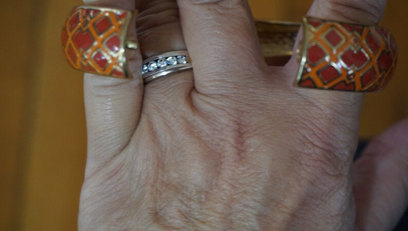 May include: A hand wearing a ring with clear stones and two orange and gold bracelets with a geometric pattern.