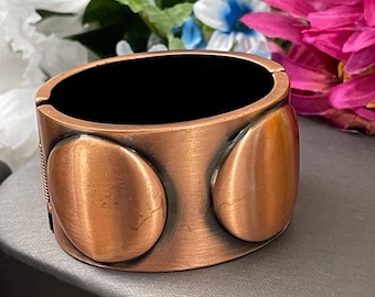 Puede incluir: Una pulsera de puño de color cobre con interior negro. La pulsera presenta tres formas ovaladas en relieve en la parte delantera y un diseño texturizado en el lateral. Una pieza de joyería llamativa.