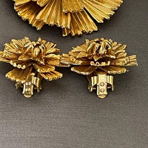 K&ouml;nnte beinhalten: Ein Set aus goldfarbenem Schmuck mit einer gro&szlig;en Brosche und einem Paar passender Ohrringe. Die Brosche und die Ohrringe bestehen aus d&uuml;nnen, filigranen Metallstreifen, die in einer blumenartigen Form angeordnet sind.