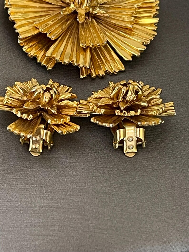 K&ouml;nnte beinhalten: Ein Set aus goldfarbenem Schmuck mit einer gro&szlig;en Brosche und einem Paar passender Ohrringe. Die Brosche und die Ohrringe bestehen aus d&uuml;nnen, filigranen Metallstreifen, die in einer blumenartigen Form angeordnet sind.